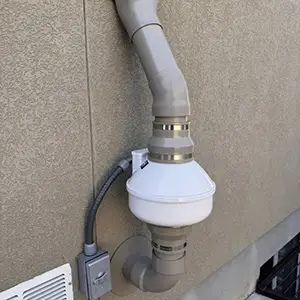 Close-up of an exterior radon fan installation: sealed pipe couplings, conduit routed flat against the wall, and a service disconnect switch within reach